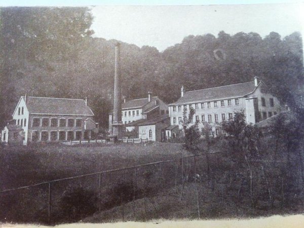 Photo en noir et blanc de l'usine l'épée. Il y a 3 bâtiments et une cheminée industrielle en lisière de forêt